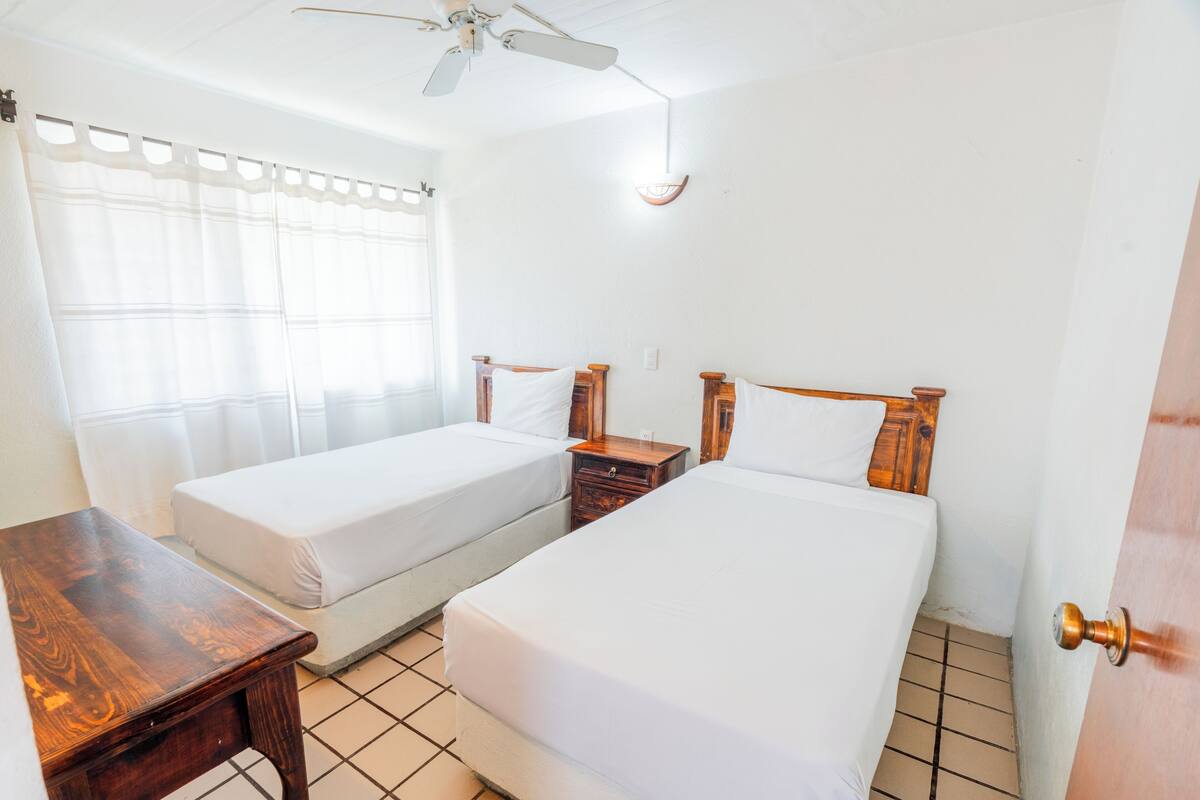 A room with two single beds, a wooden nightstand, a ceiling fan, and a window with sheer curtains.
