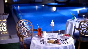 A dining table set with a candle, a glass of red drink, and plates of food, overlooking a pool. - Corp Executive Hotel Doha Suites (Doha)