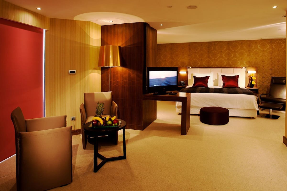 A hotel room with a bed, a TV, a chair, a small table with fruit, and a trash bin.