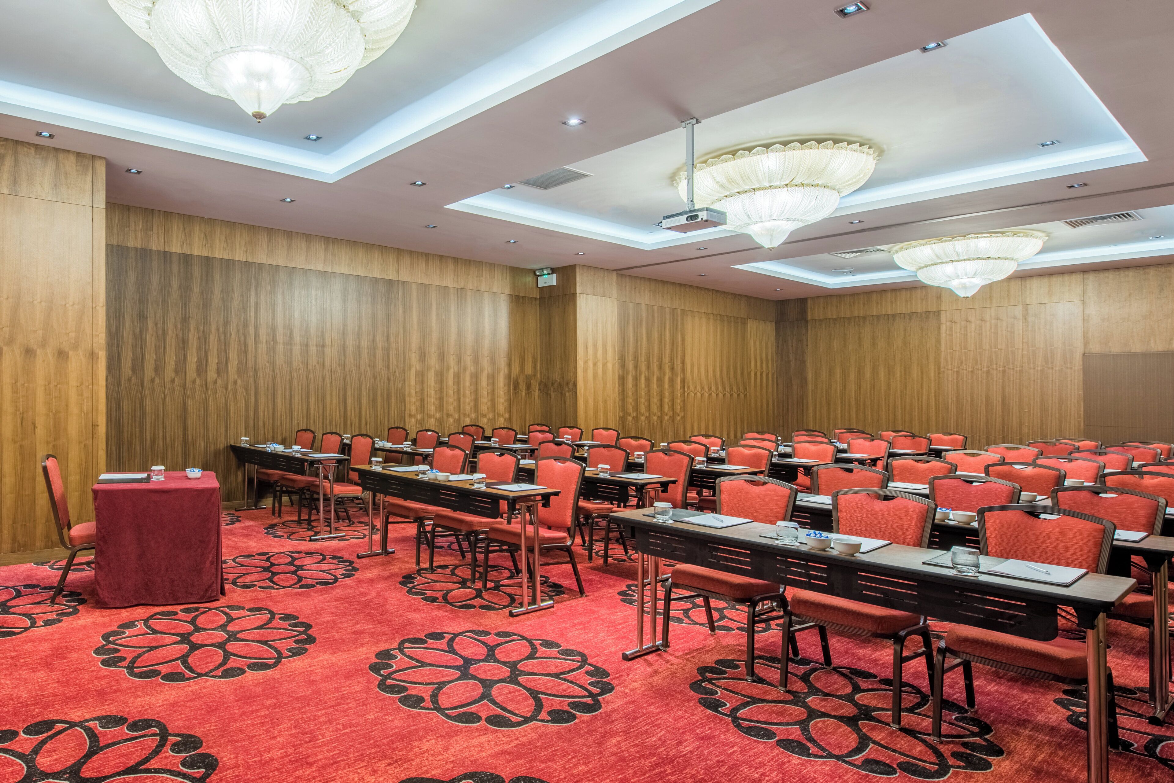 Uma sala de conferências com fileiras de mesas e cadeiras, paredes de painel de madeira e luminárias decorativas no teto.