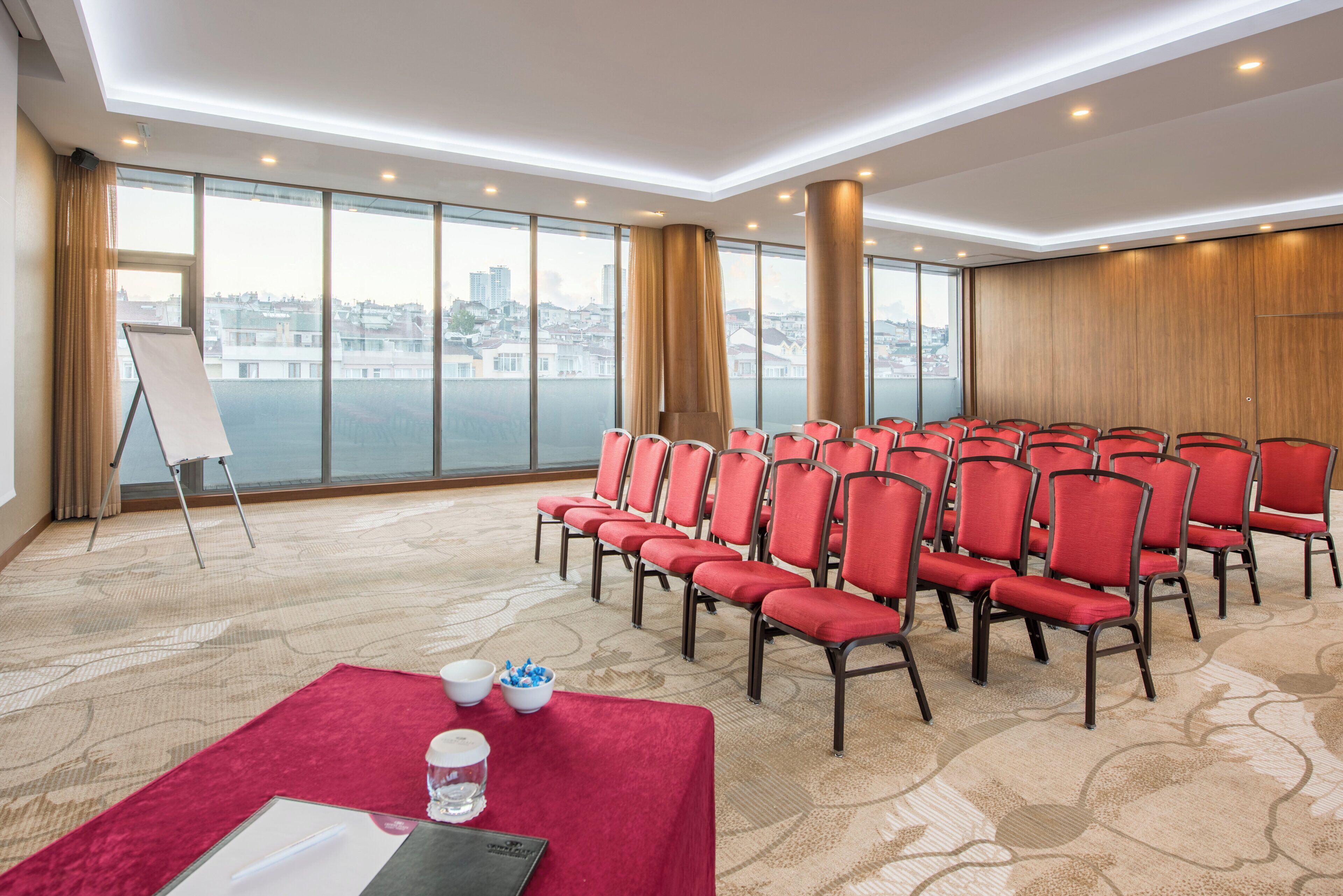 A conference room with rows of red chairs, a whiteboard, and a large window overlooking a cityscape.