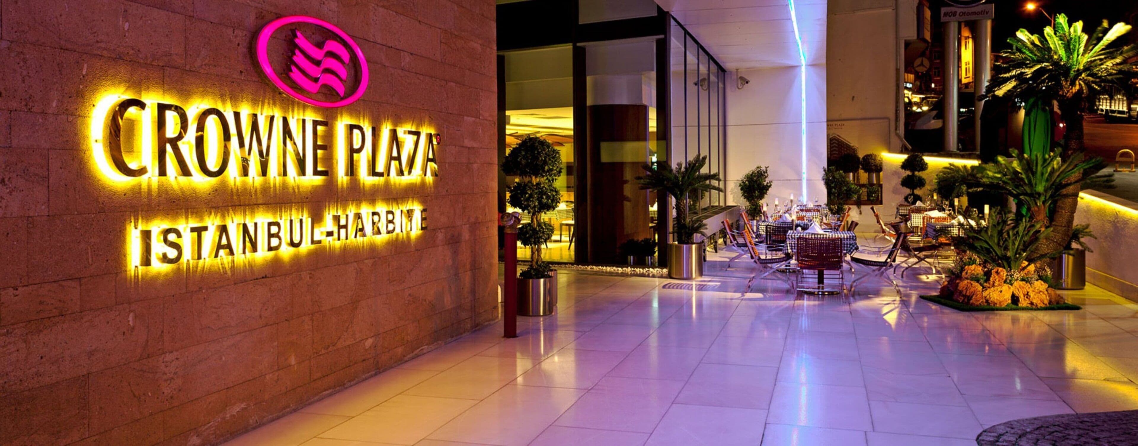 A hotel entrance with a neon sign for Crowne Plaza Istanbul-Harbiye, outdoor seating area, and potted plants.