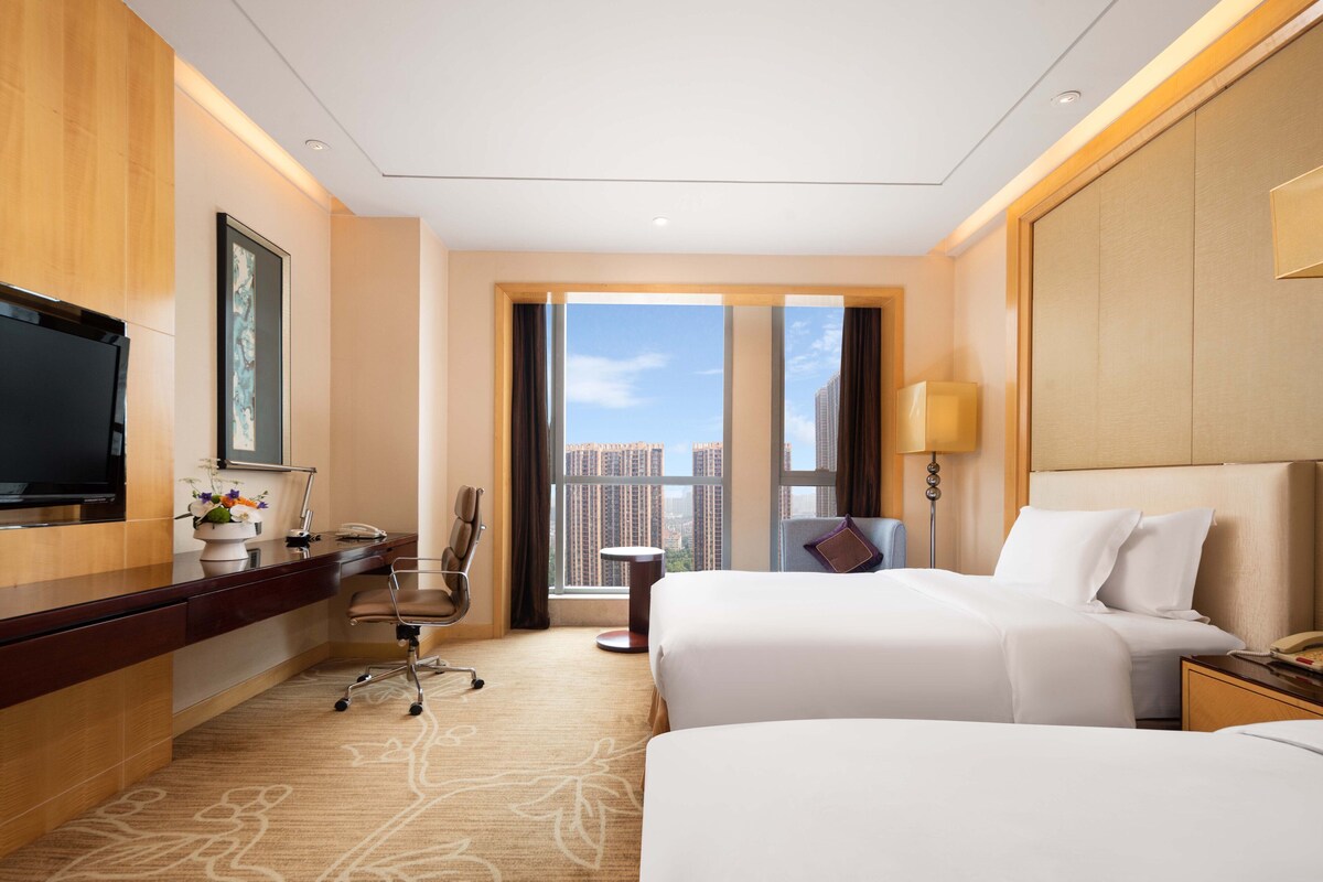 A hotel room with a large bed, a desk with a computer, a chair, a TV, and a view of the city.
