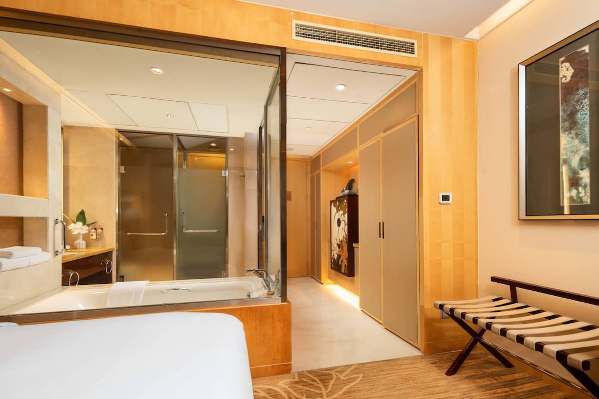 A modern hotel room with a large bed, a separate bathroom with a glass shower, a bench, and a painting on the wall.