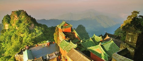 Un complejo arquitectónico tradicional chino con varios edificios, un patio interior y un paisaje montañoso de fondo.