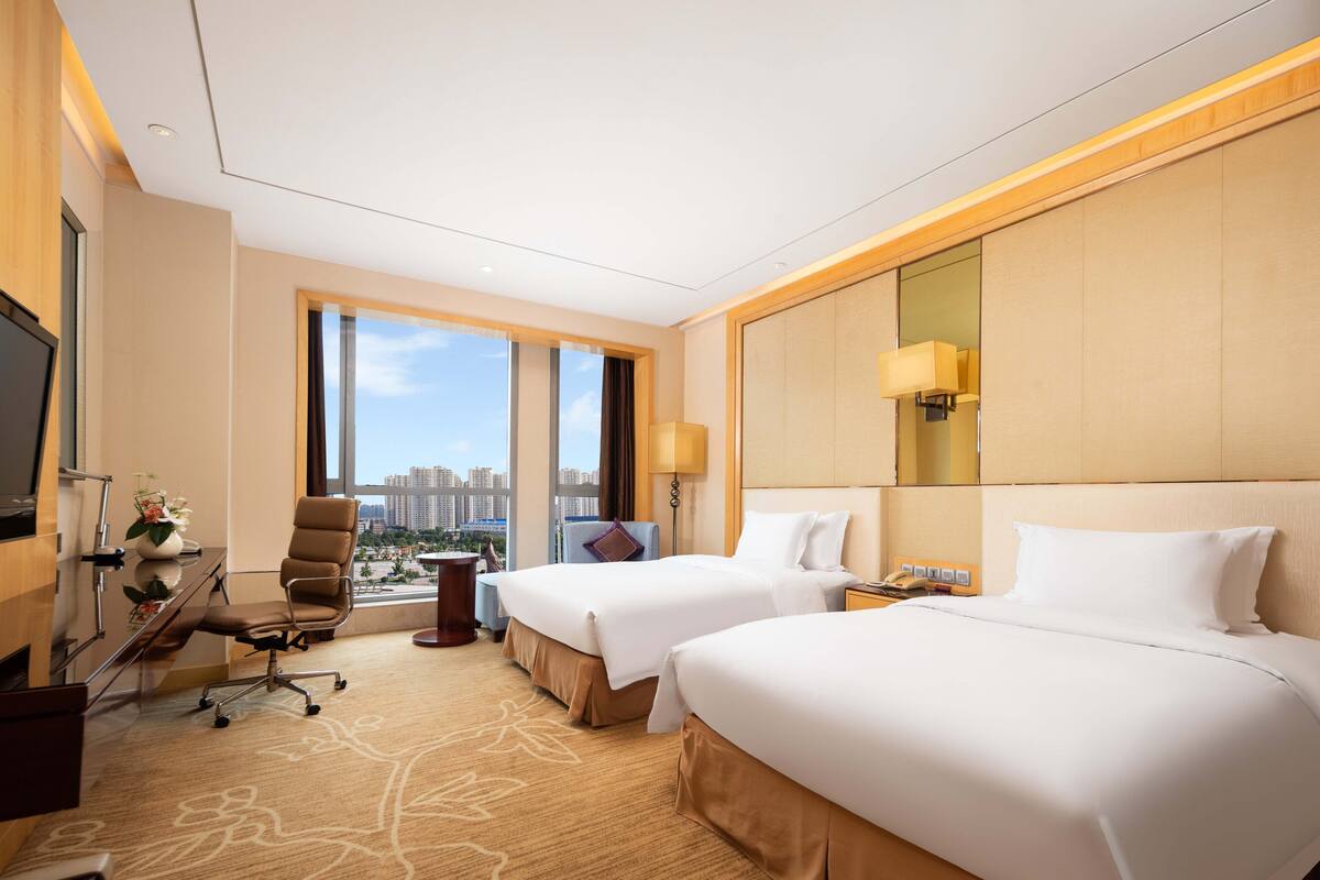 A hotel room with two beds, a desk with a chair, a TV, and a large window offering a city view.