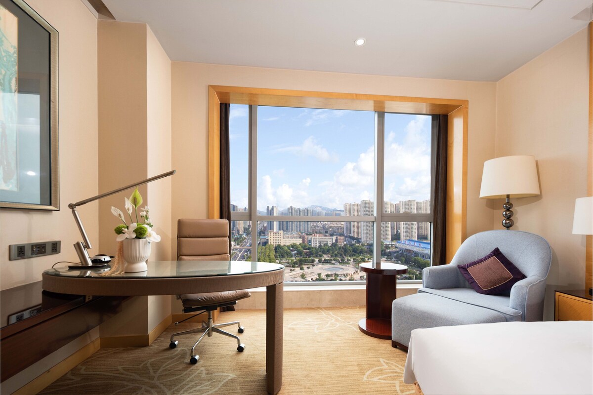 A hotel room with a large window, a desk with a lamp, a chair, and a bed.