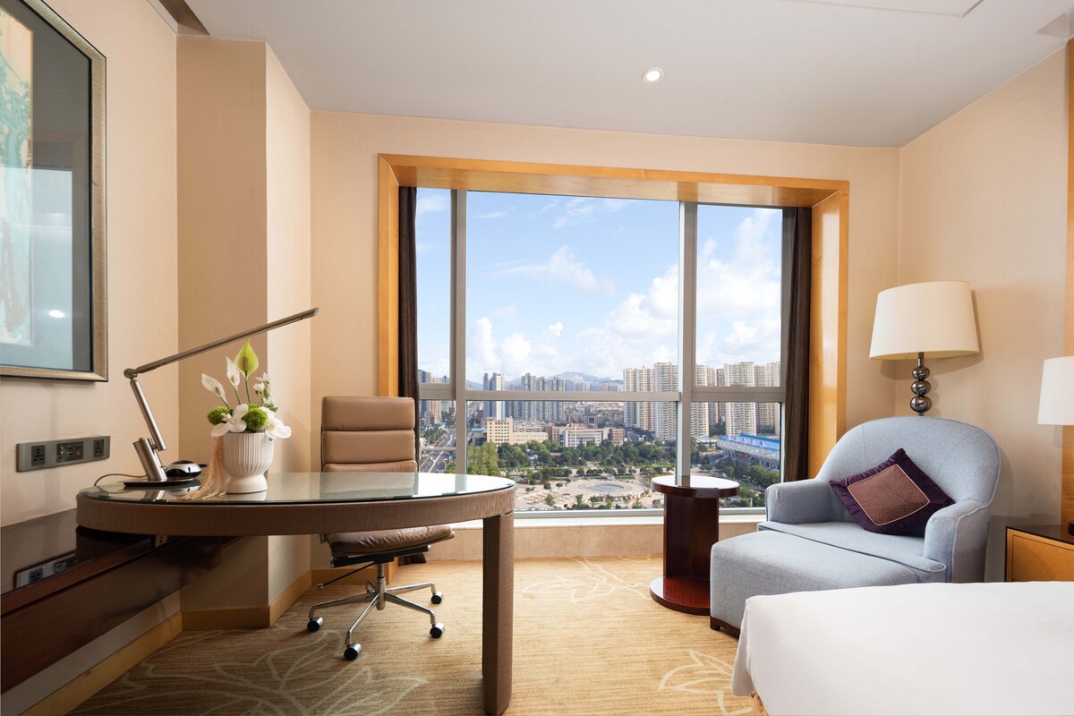 A hotel room with a large window, a desk with a lamp, a chair, and a bed.