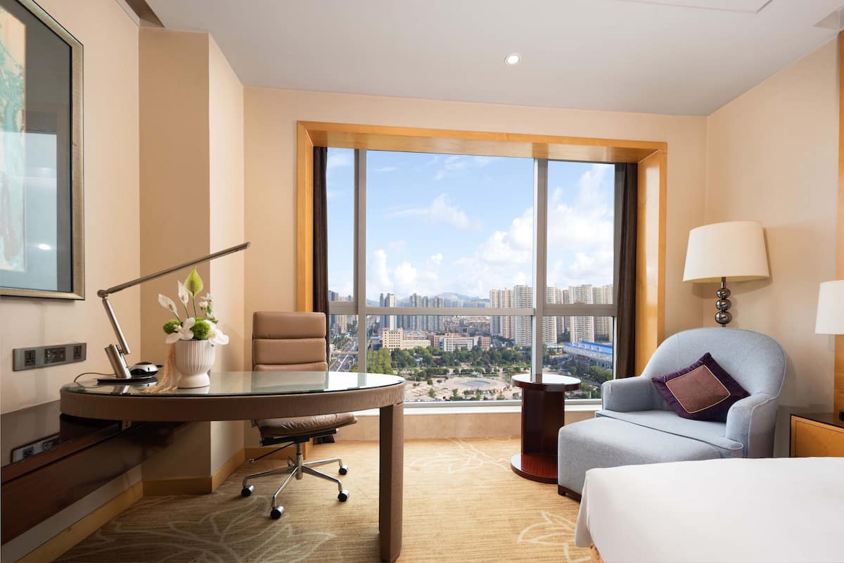 A hotel room with a large window, a desk with a lamp, a chair, and a bed.