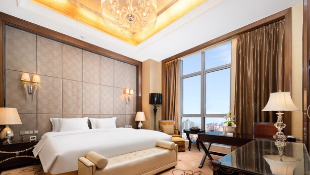 Amplia habitación de hotel con una cama grande, un escritorio, una silla y vistas de la ciudad a través de amplios ventanales.