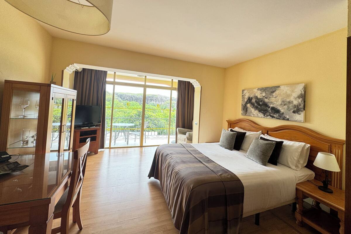 A hotel room with a bed, a desk with a TV, a balcony with a view of greenery, and a painting on the wall.