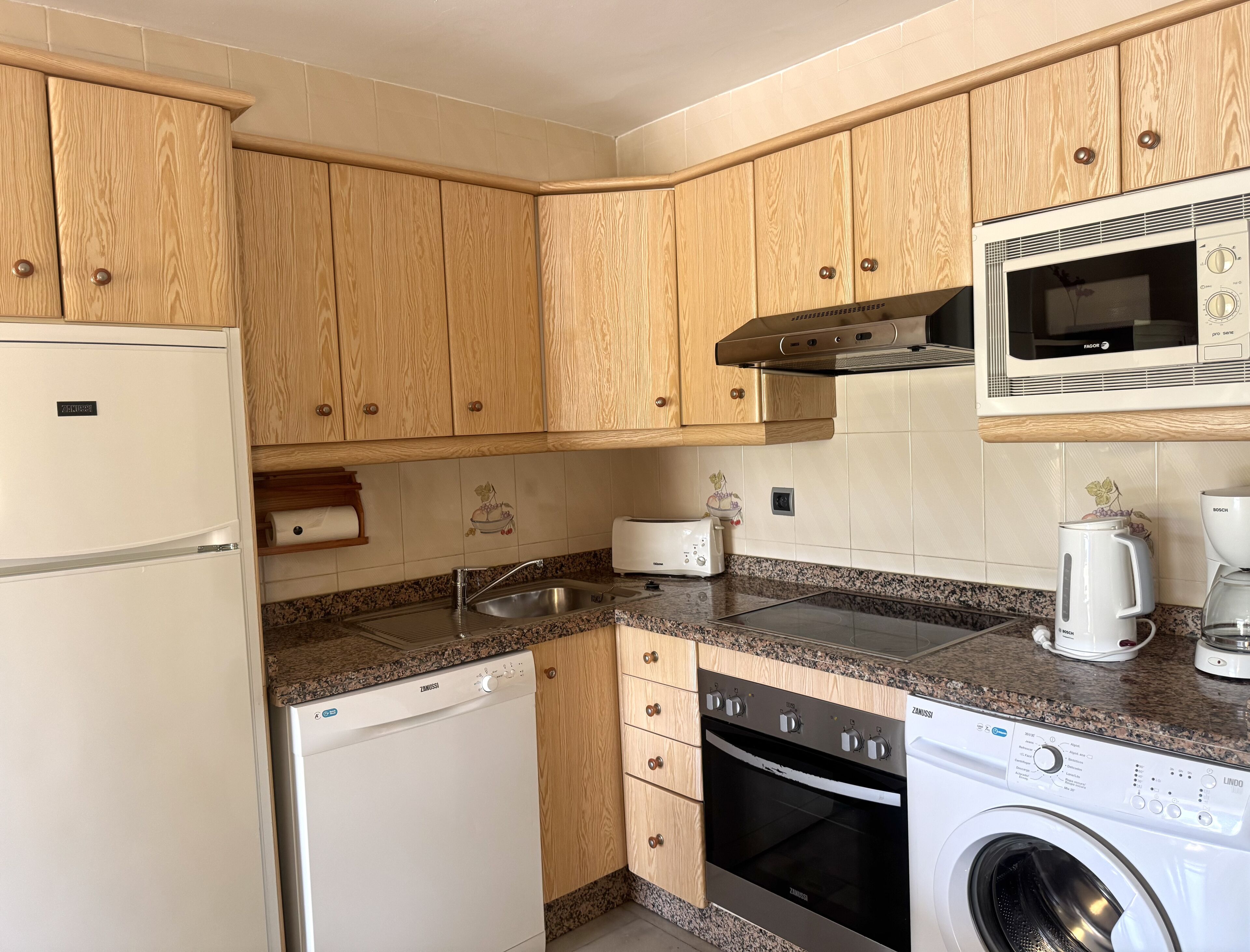 A compact kitchen with wooden cabinets, a microwave, a dishwasher, an oven, and a washing machine.