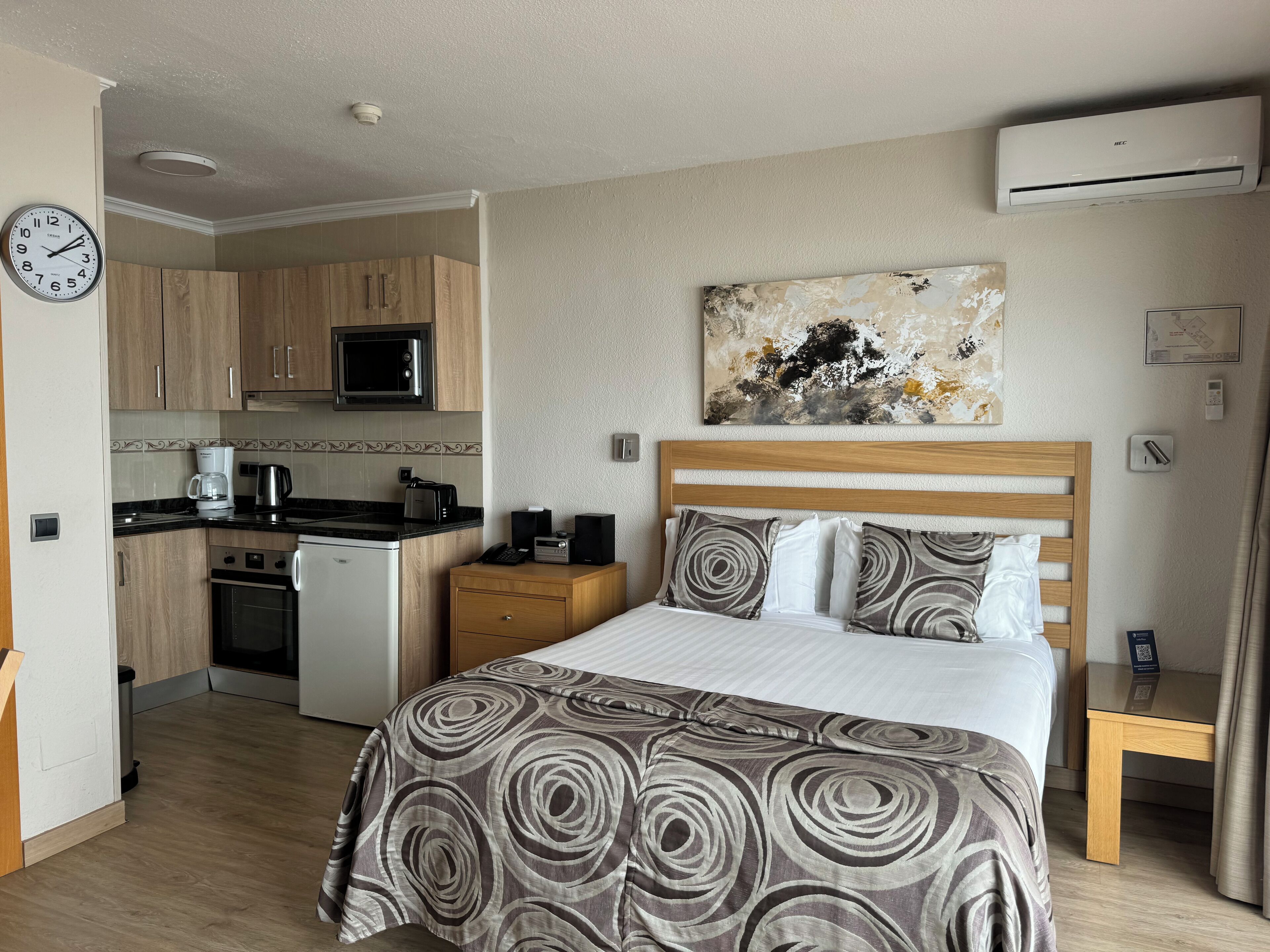 Et kompakt hotelværelse med tekøkken, en seng med mønstret sengetøj, et billede på væggen og en loftmonteret aircondition.