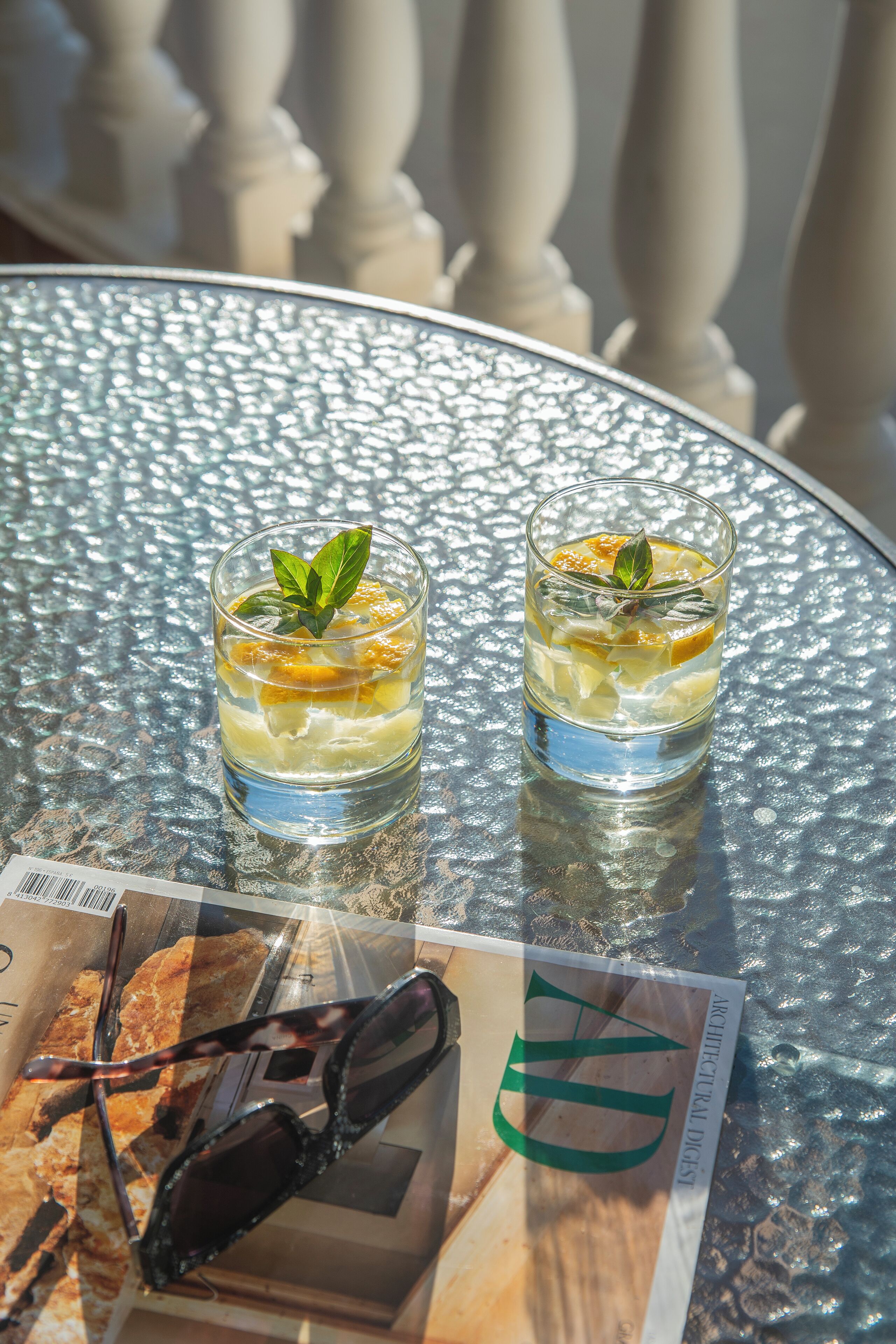 Two glasses of a beverage with a mint leaf garnish on a glass table.