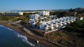 A coastal hotel with multiple white buildings, a beach with umbrellas, and a view of the ocean and mountains in the background.