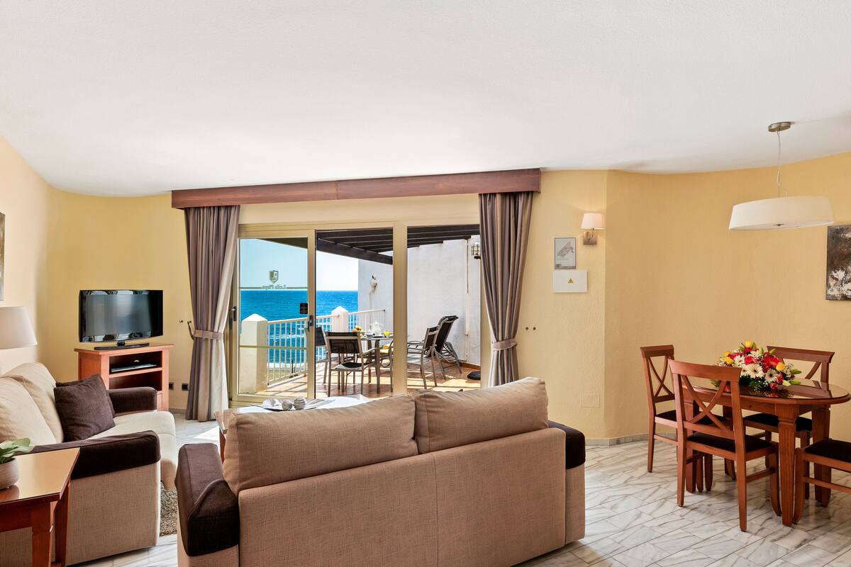 A hotel room with a sofa, TV, dining table, and chairs. There is a balcony with a view of the sea.