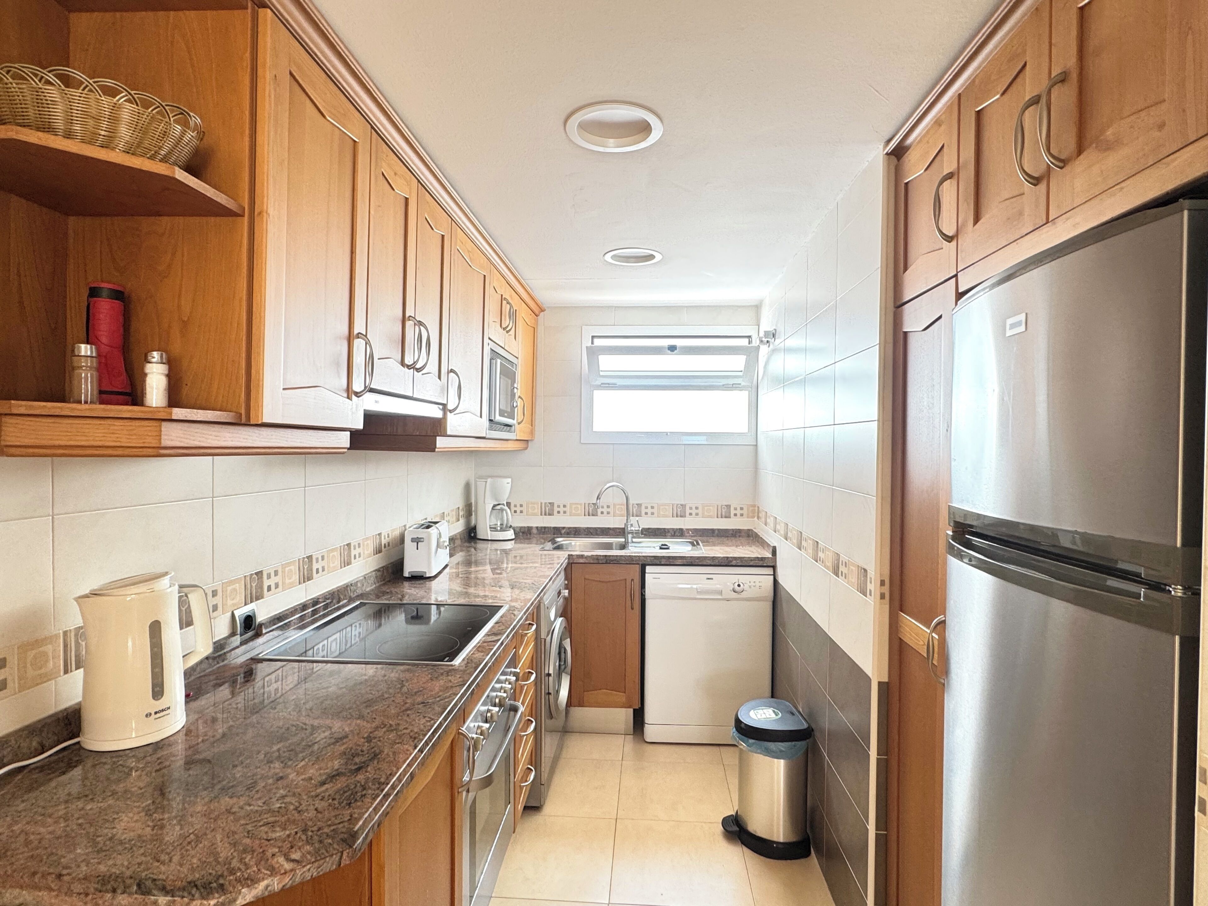A compact kitchen with wooden cabinets, a stainless steel refrigerator, a built-in oven, a sink, and a dishwasher.