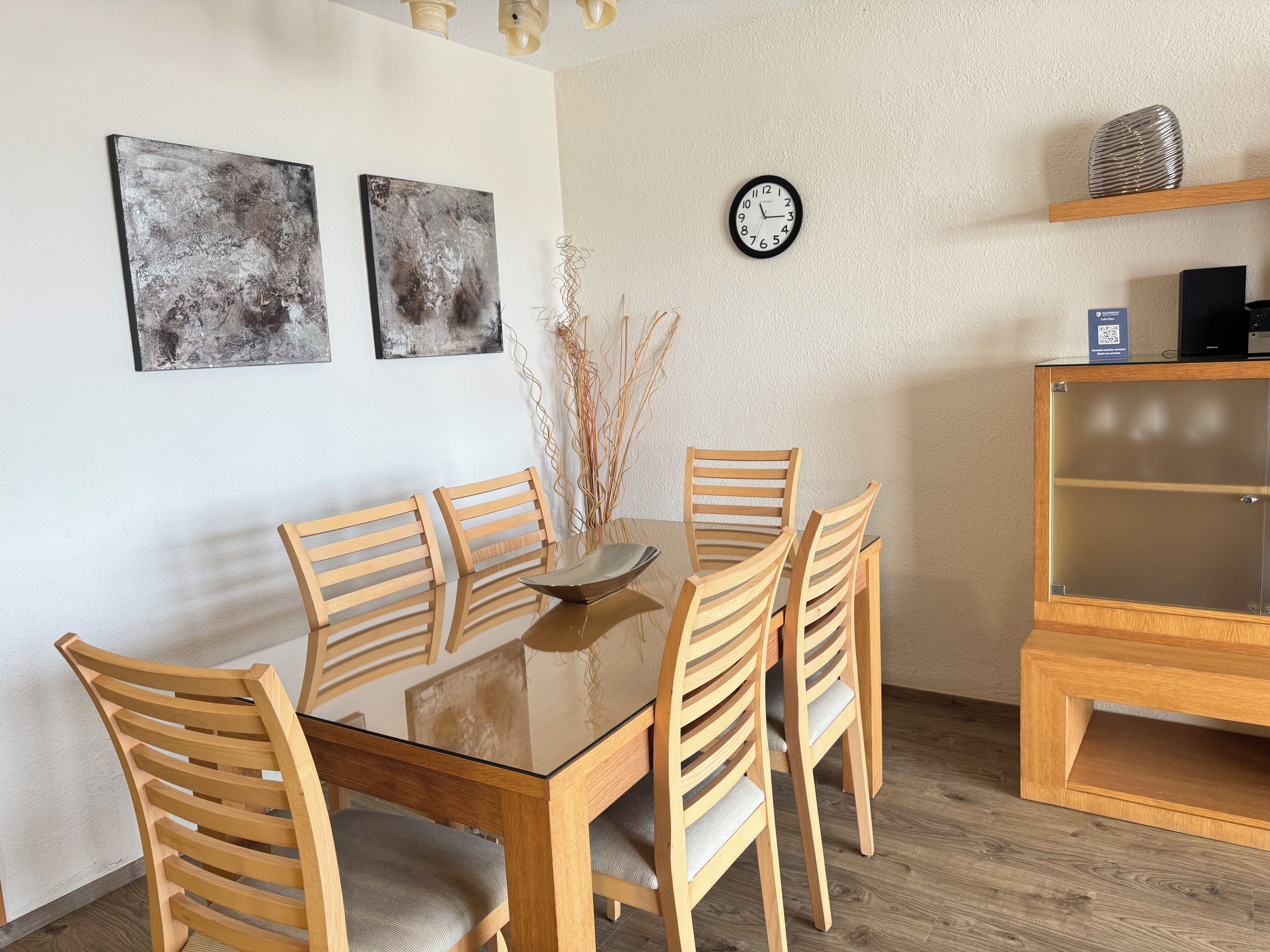 A dining area with a glass-top table surrounded by wooden chairs, two abstract paintings on the wall, a clock, and a wooden cabinet with a QR code.
