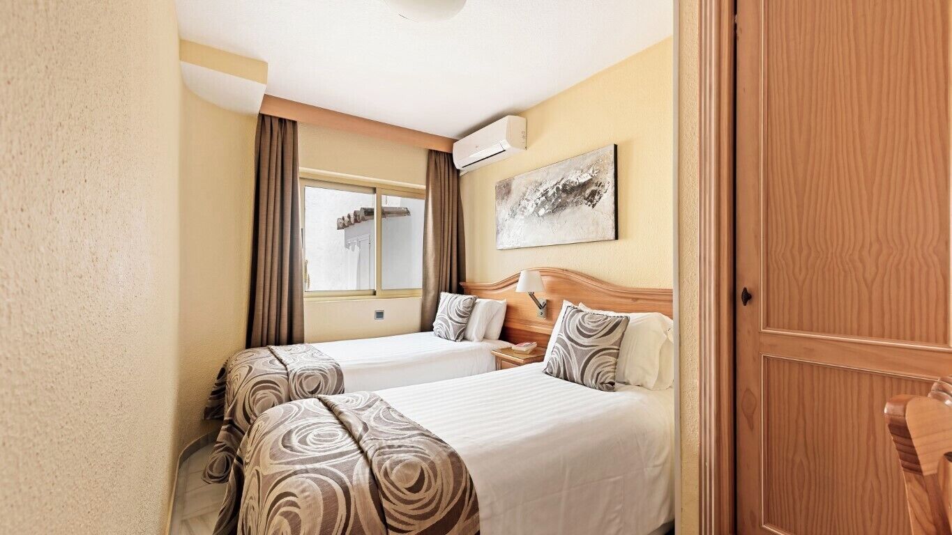 A hotel room with two beds, a window, an air conditioning unit, and a wooden door.