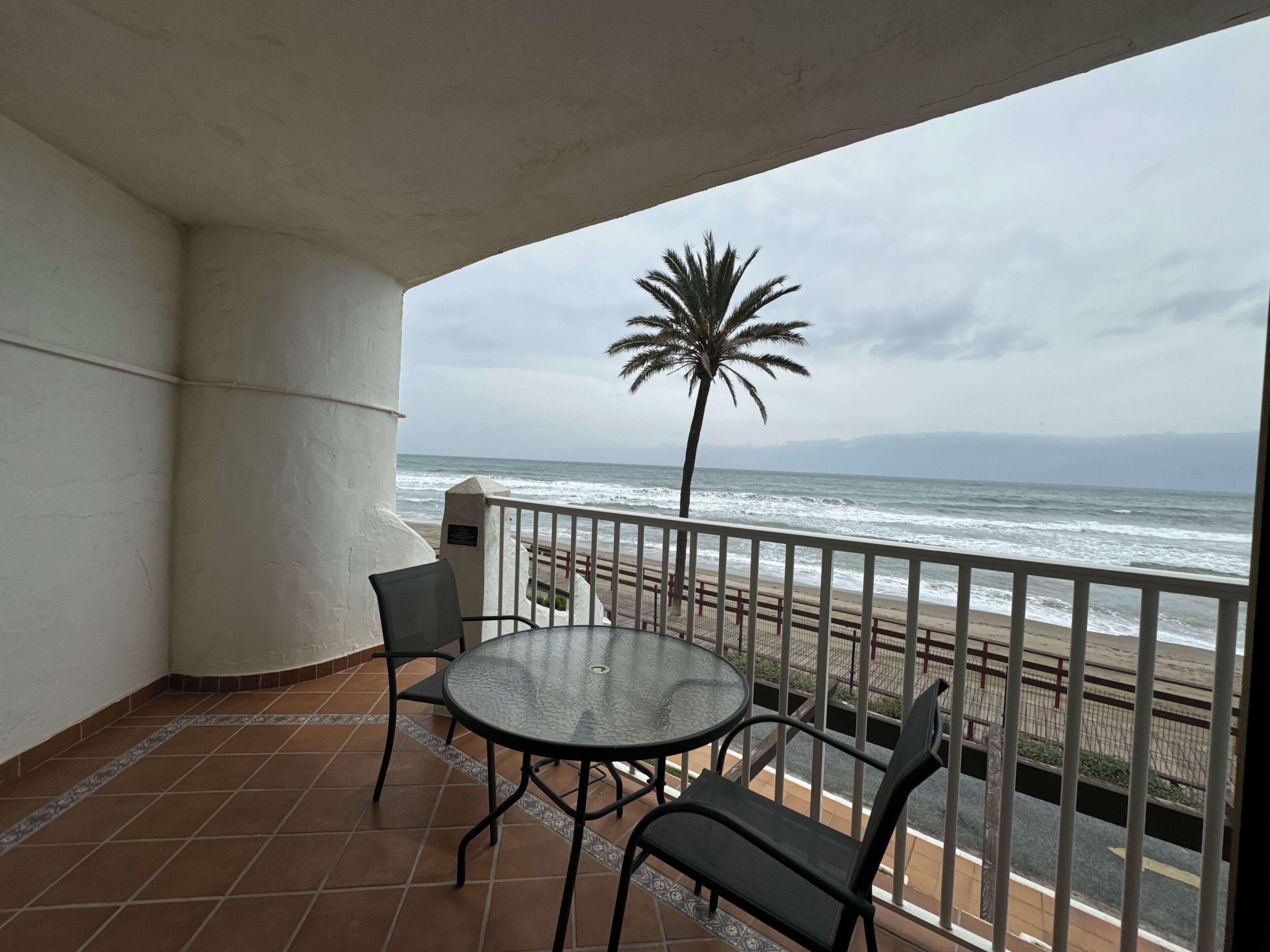 Un balcón con una mesa redonda de vidrio y dos sillas, con vistas a una playa y al océano, donde se distingue una palmera.