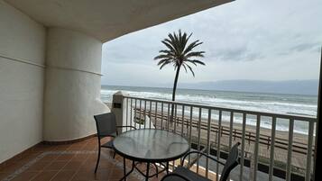 Un balcón con una mesa redonda de vidrio y dos sillas, con vistas a una playa y al océano, donde se distingue una palmera.