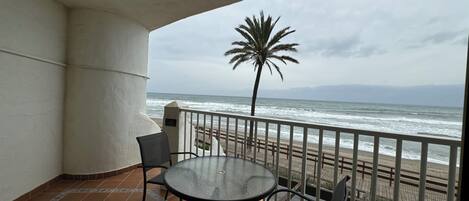 A balcony with a round glass table and two chairs, overlooking a beach with a palm tree and ocean view.