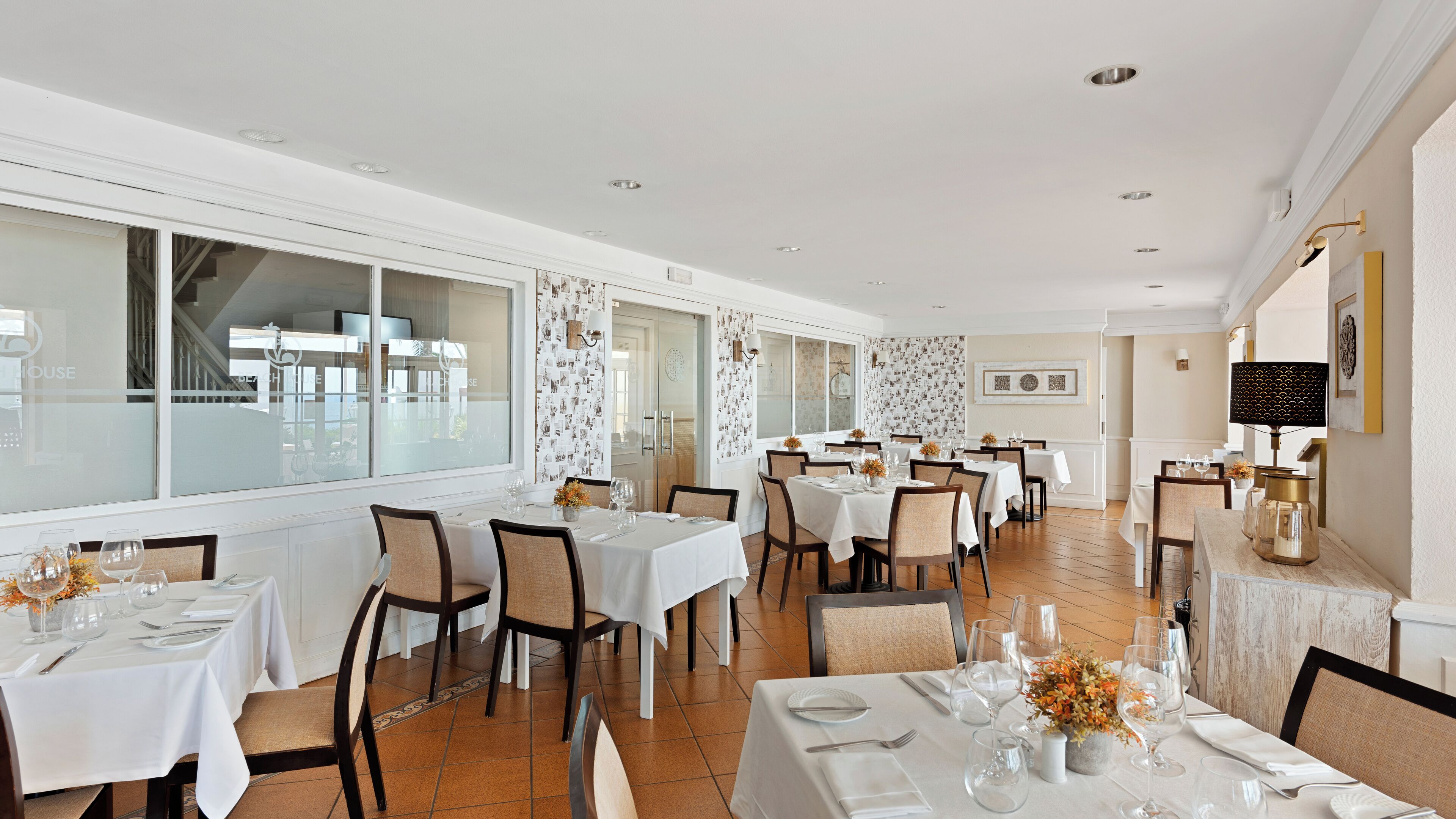 A spacious dining area with multiple tables set for dining, large windows providing natural light, and a view of the outside.