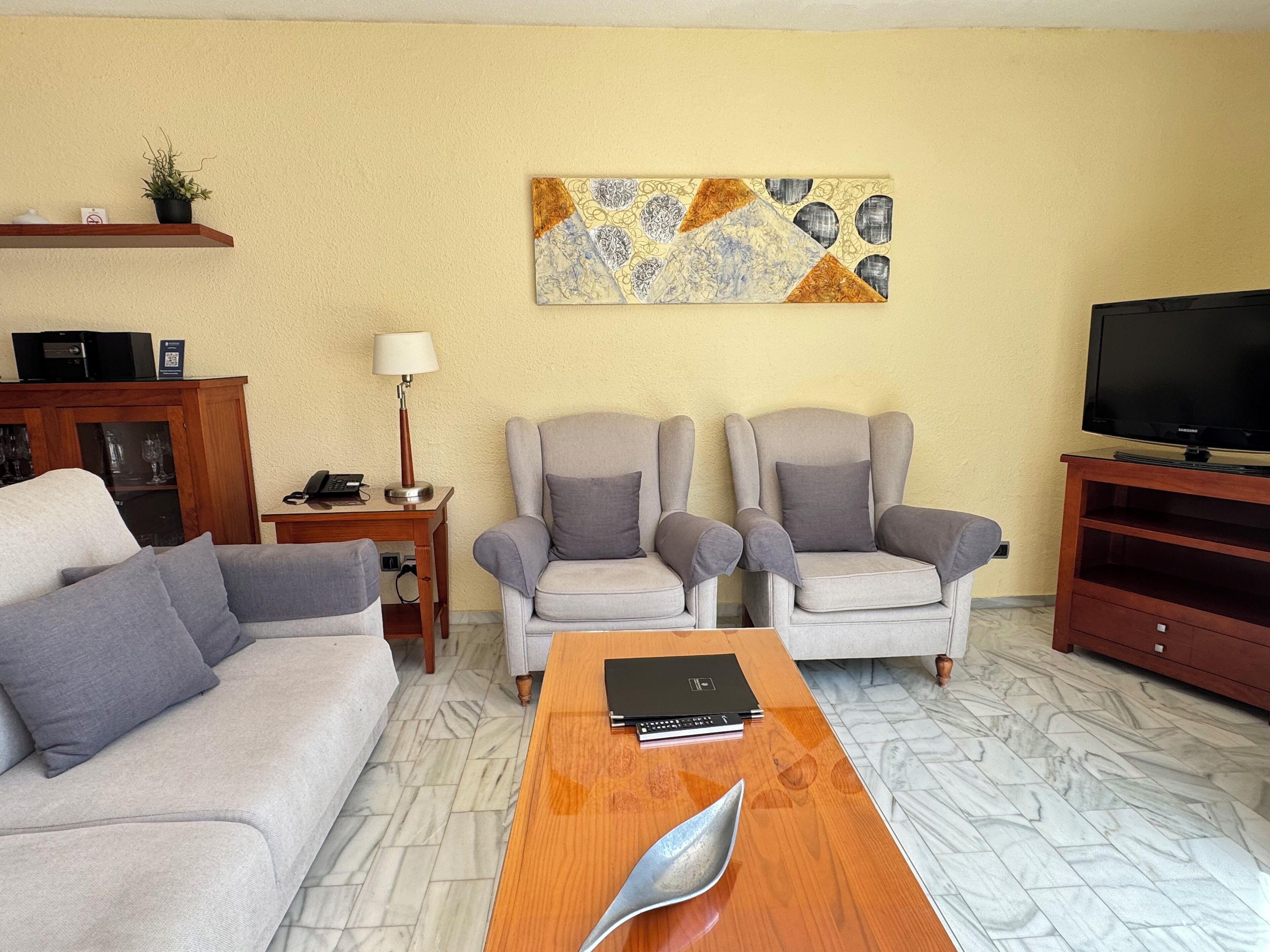 Una sala de estar con un sofá, dos sillones, una mesita de centro, un televisor, una lámpara y un adorno en la pared.