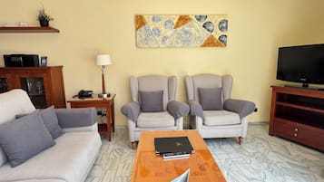 Una sala de estar con un sofá, dos sillones, una mesita de centro, un televisor, una lámpara y un adorno en la pared.