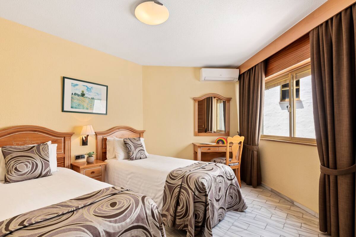 A hotel room with two beds, a desk, a chair, a window with curtains, a ceiling light, and a painting on the wall.
