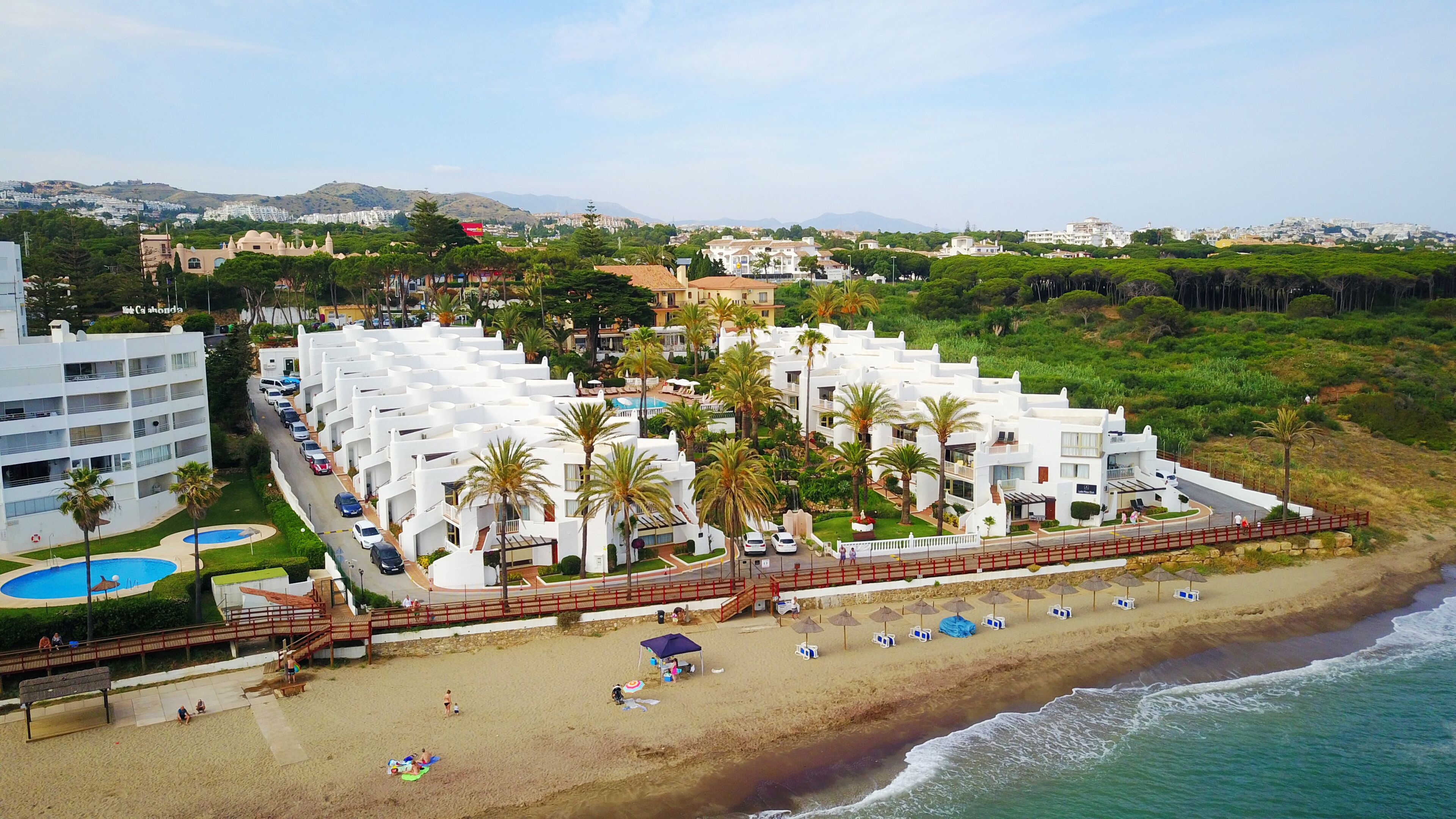 Un hotel costero con edificios blancos, palmeras, una playa y vistas al mar.