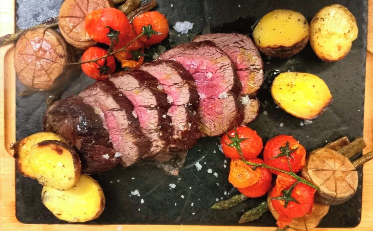 A steak with roasted potatoes and tomatoes on a slate board.