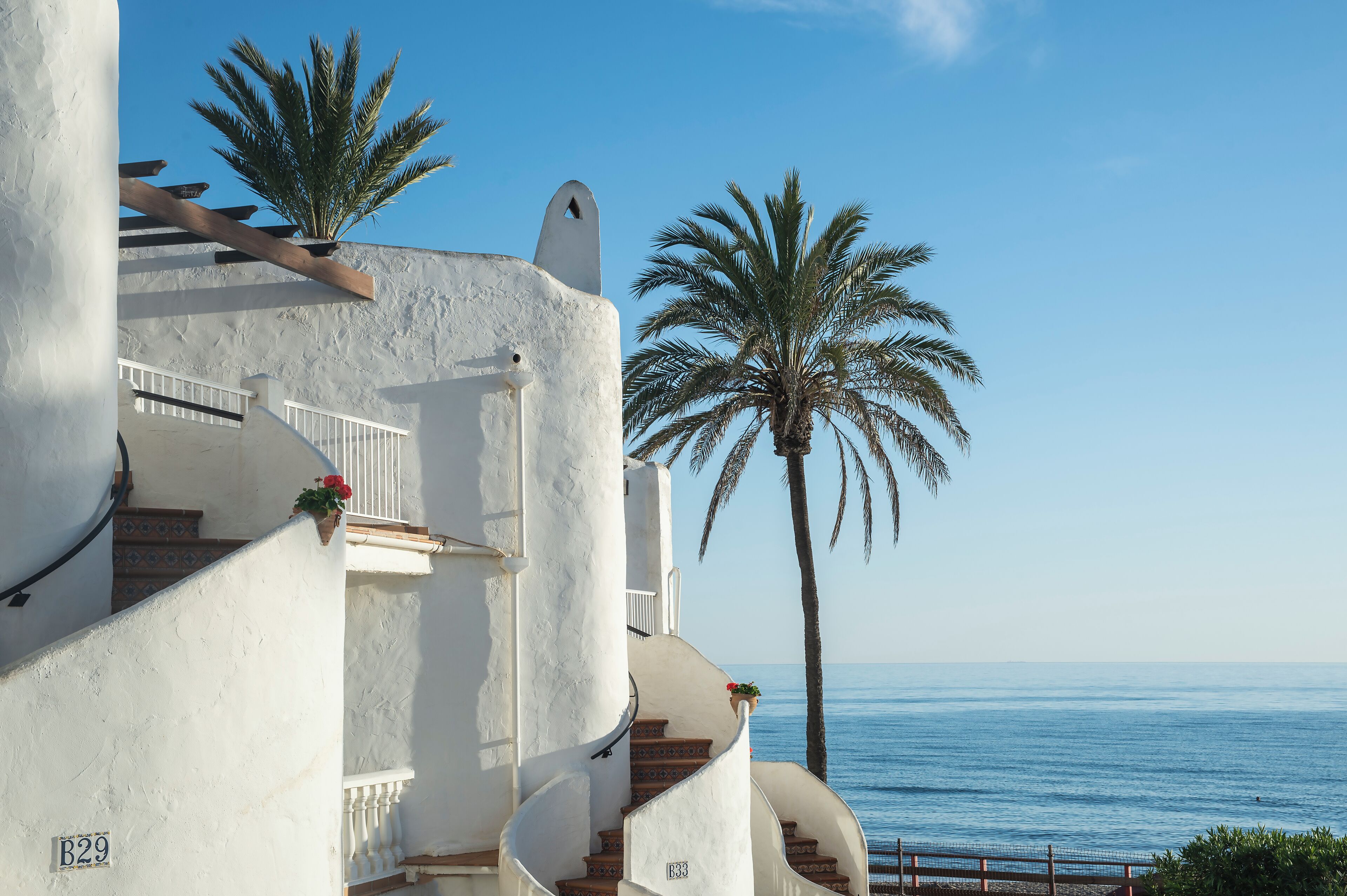 A white building with a spiral staircase, palm trees, and a view of the sea.