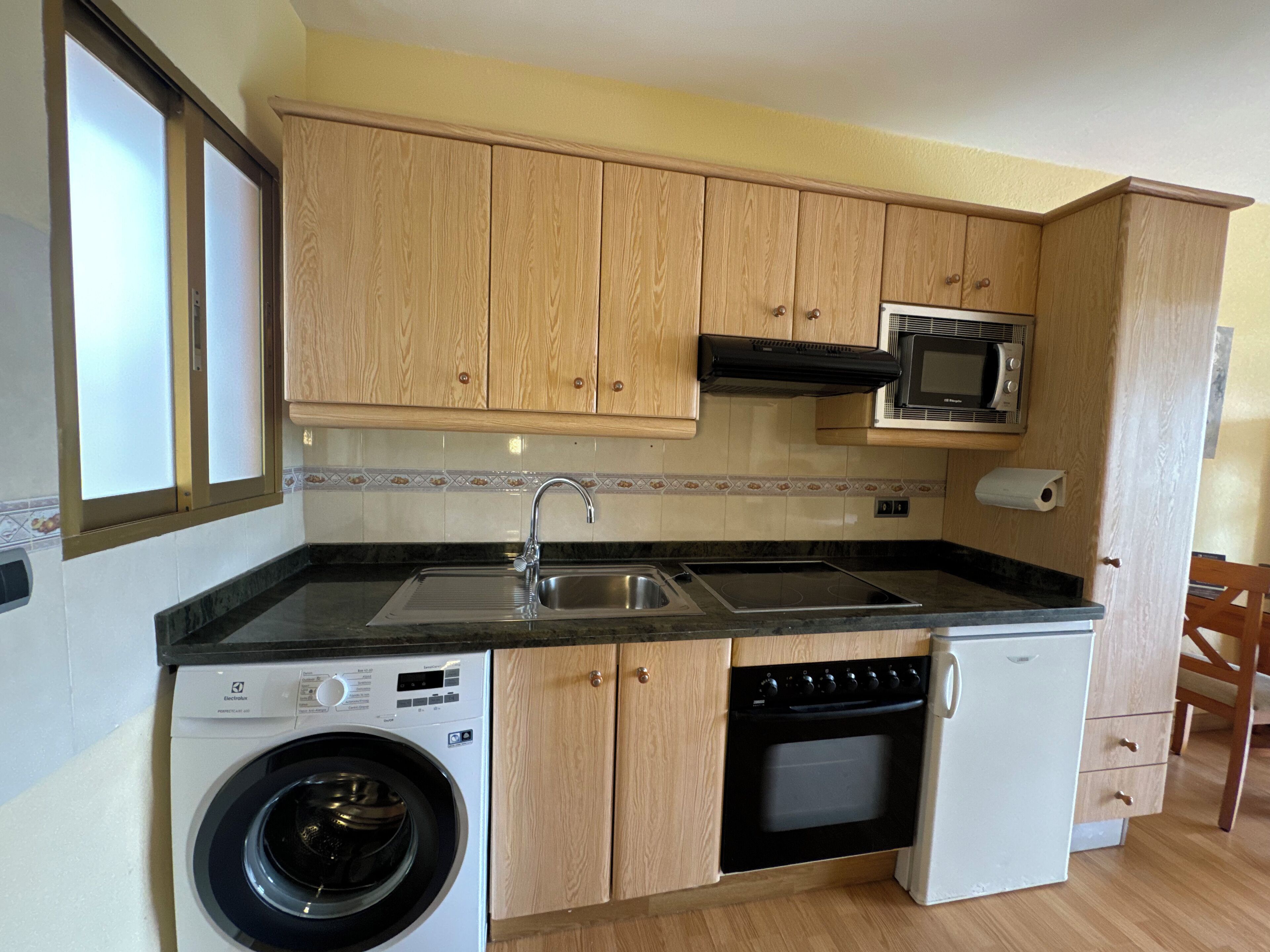 A compact kitchen with wooden cabinets, a built-in microwave, a washing machine, and a dishwasher.