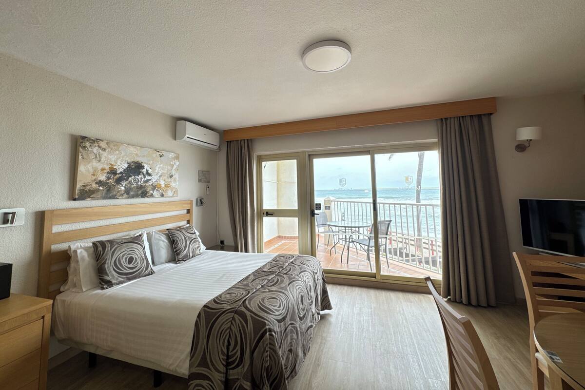 A hotel room with a bed, a TV, a balcony with a view of the sea, and a painting on the wall.