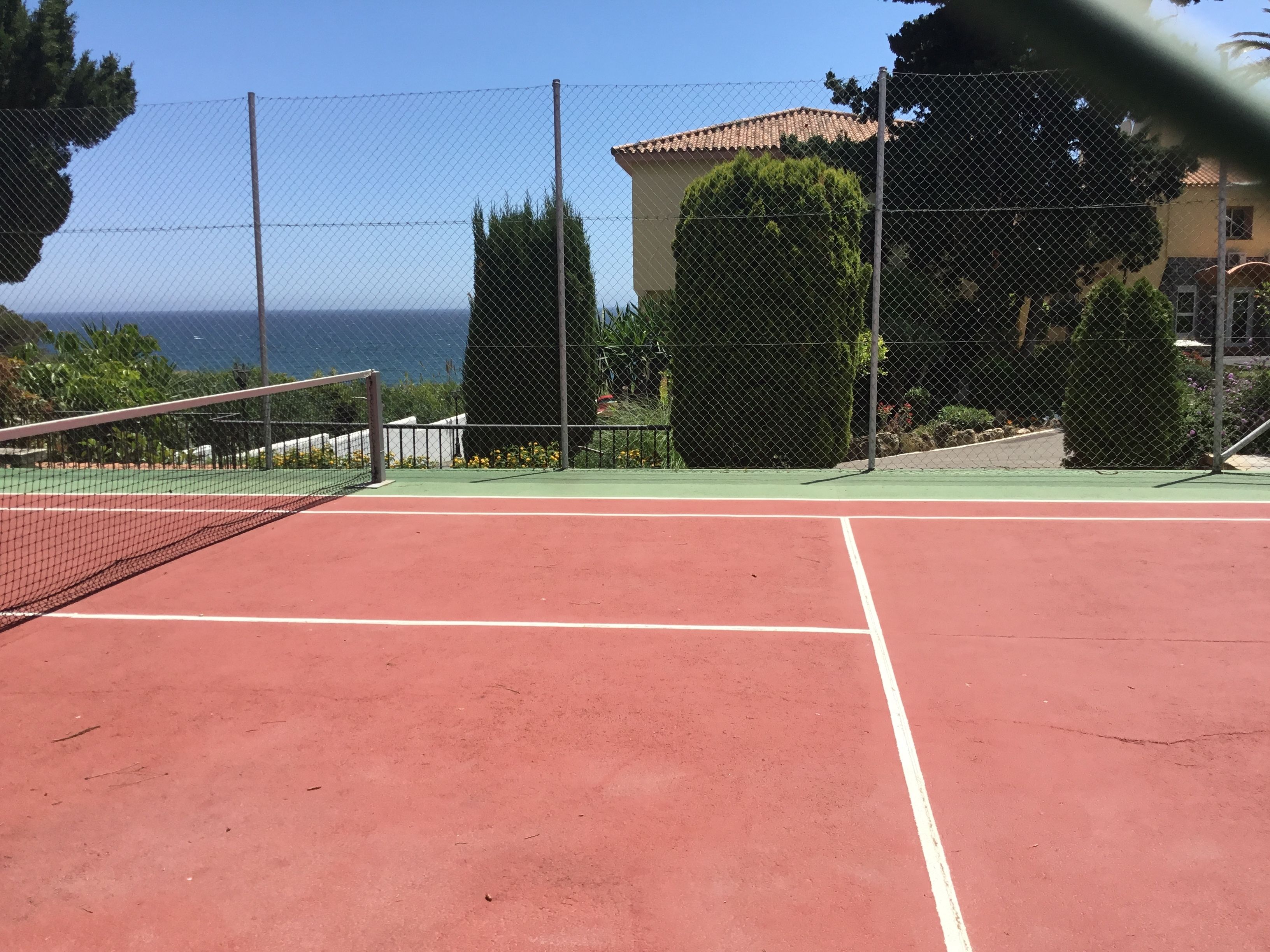 A tennis court with a net, surrounded by a chain-link fence and greenery.