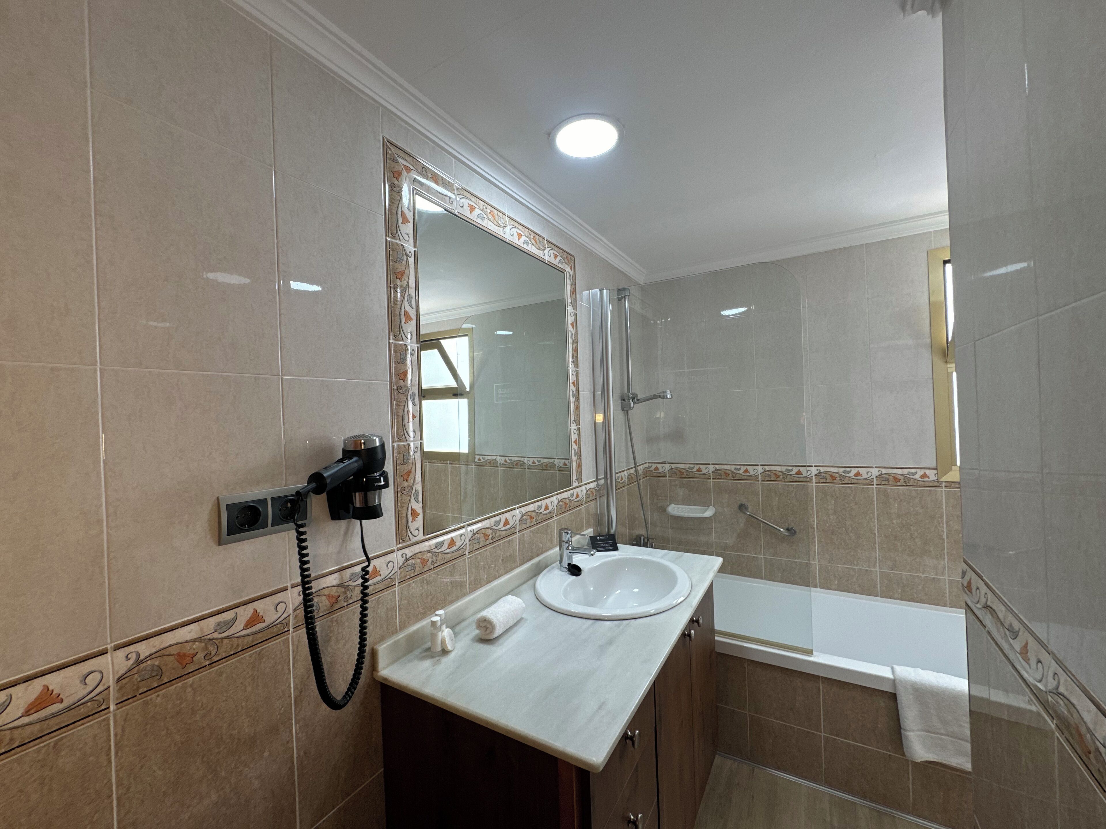 A bathroom with a sink, mirror, shower, and a hairdryer on the wall.