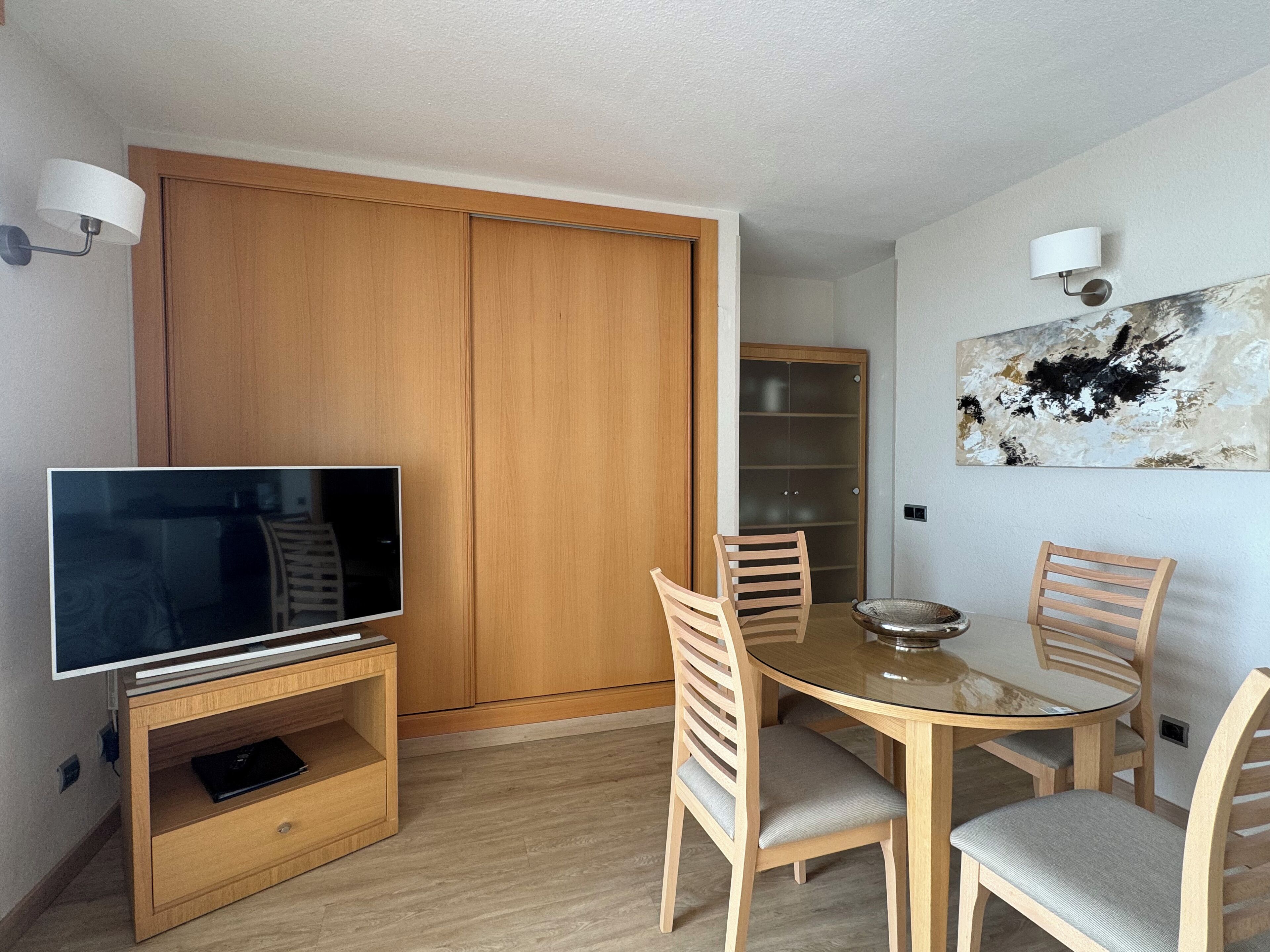 A compact living space with a TV on a wooden stand, a dining table with chairs, a wooden wardrobe, and a wall-mounted abstract painting.