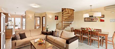 A spacious living room with a large beige sectional sofa, a glass coffee table, and a wooden dining table with chairs. A wooden spiral staircase is visible in the background.