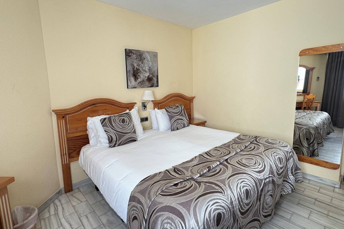 A hotel room with a large bed, a bedside table, a lamp, a mirror, and a wall-mounted artwork.