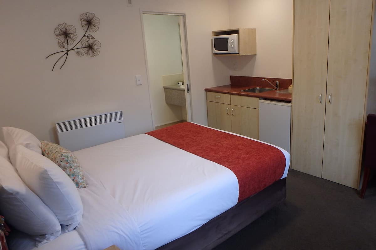 A bedroom with a bed, a red blanket, a wooden wardrobe, a small kitchenette with a microwave, and a mirror above the mirror cabinet.