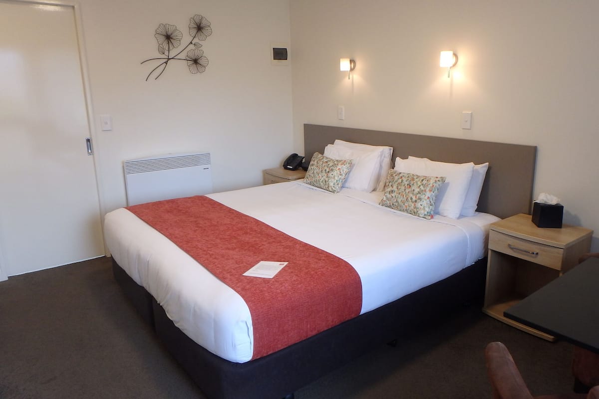 A neatly made bed with a red blanket, a bedside table with a lamp, and a chair in a hotel room.