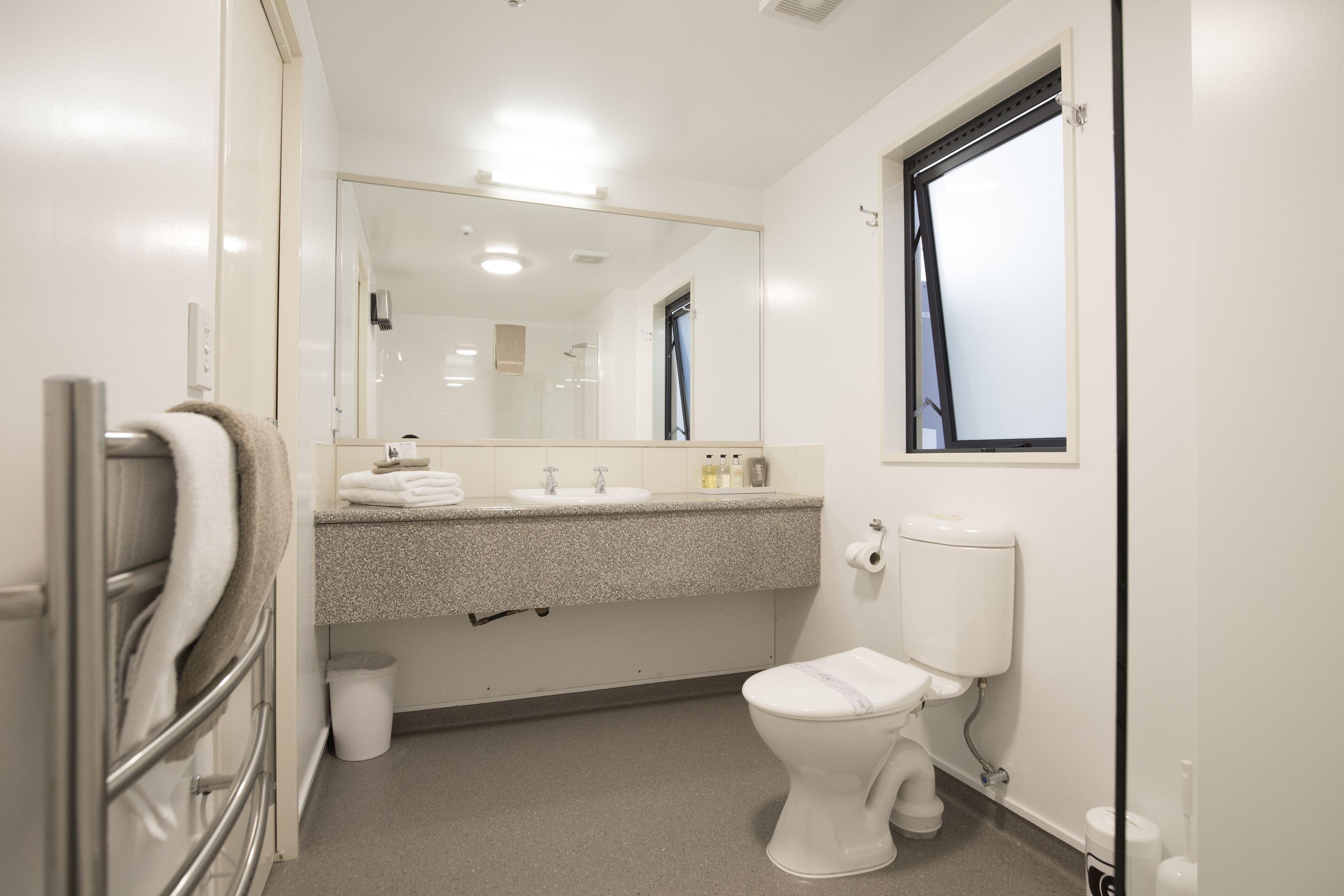 Ein modernes Badezimmer mit Toilette, Waschbecken und Spiegel. Handtuchhalter und Mülleimer sind ebenfalls vorhanden.