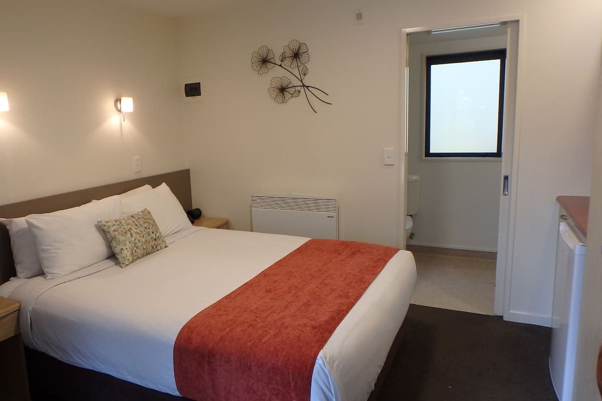 A hotel room with a bed, bedside tables, a nightstand, a wall-mounted lamp, a decorative wall piece, and a door leading to a bathroom.