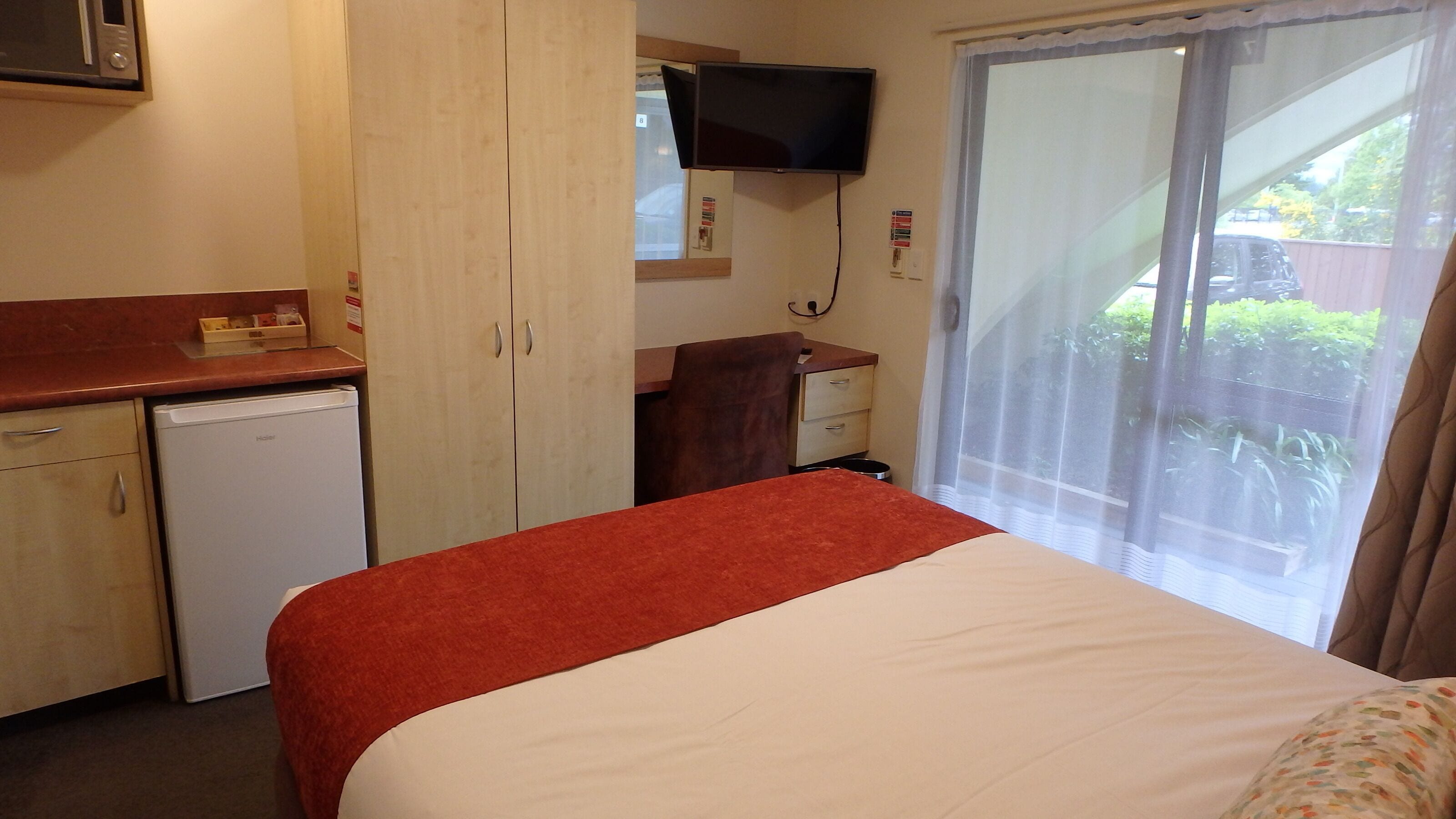 Ein Hotelzimmer mit Bett, Schreibtisch, kleinem Kühlschrank, an der Wand montiertem Fernseher und einem Fenster mit Ausblick.