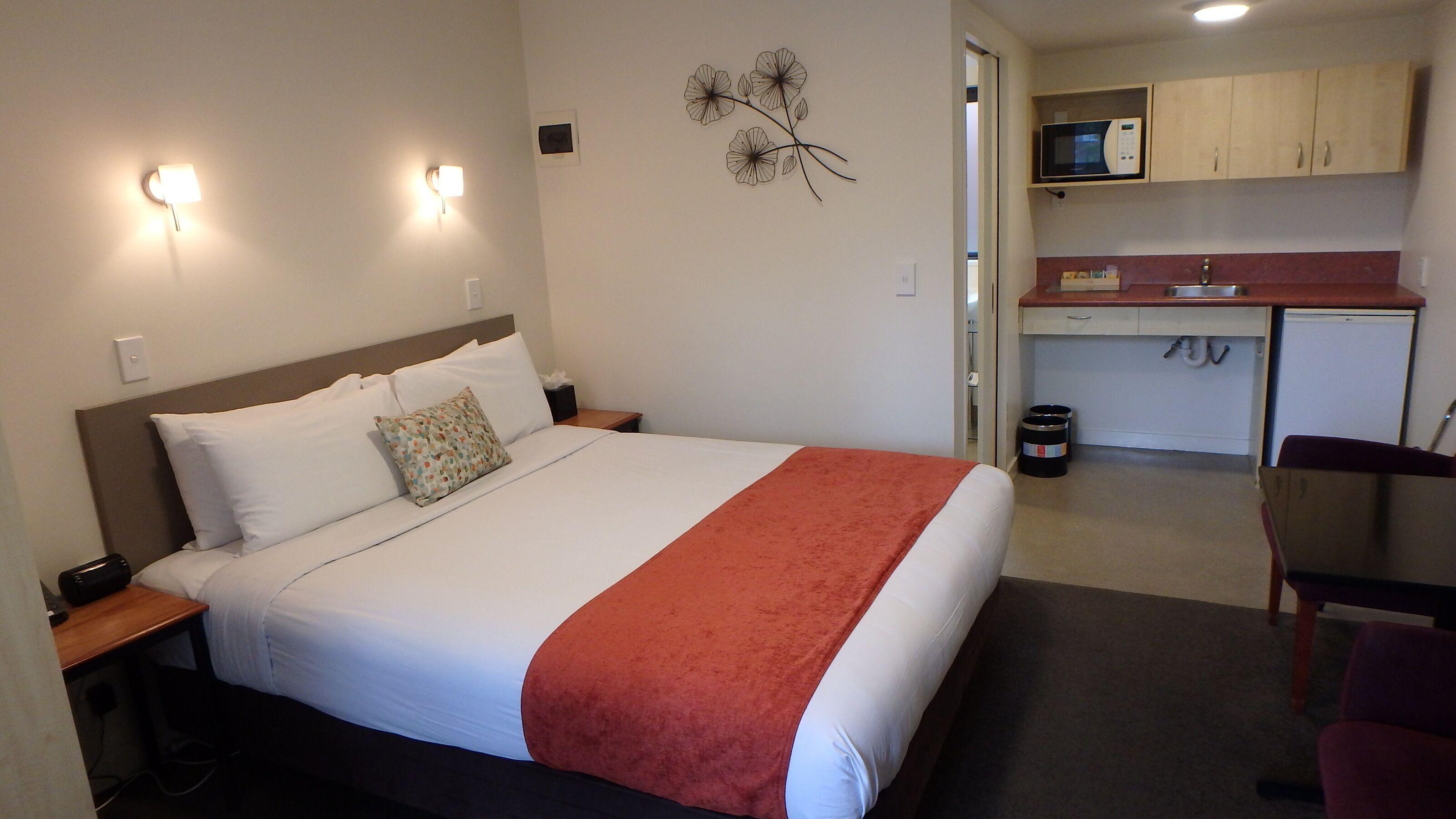 Habitación de hotel con cama, mesita de noche, lámpara y un área de kitchenette.