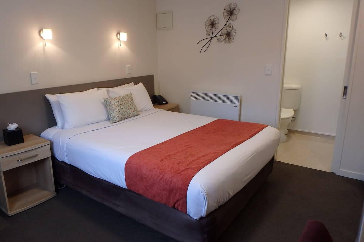 A hotel room with a bed, bedside tables, a nightstand, a lamp, a wall decoration, and a bathroom door.