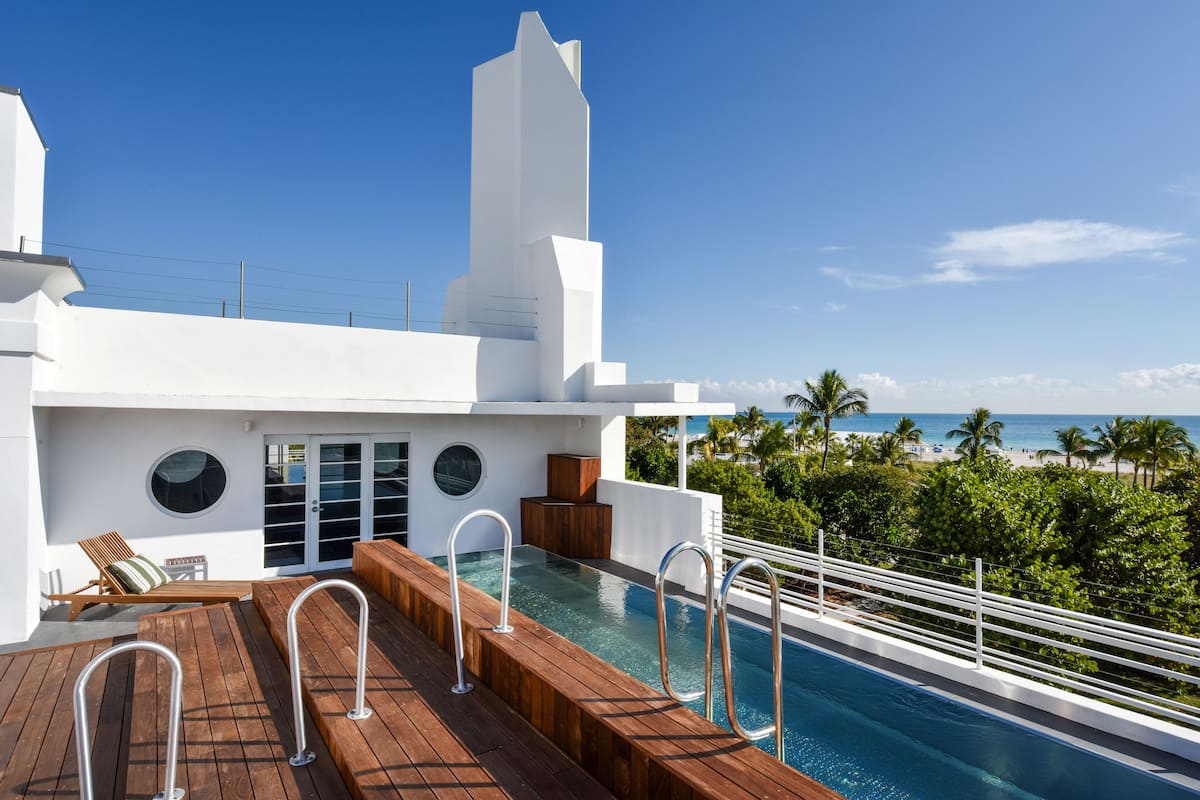 A modern outdoor pool area with wooden decking, a lounge chair, and a view of the beach and palm trees.