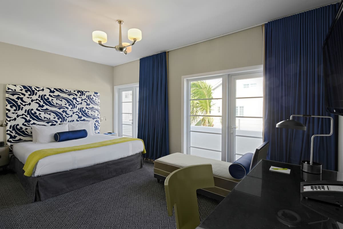 A hotel room with a bed, a desk, a chair, a desk lamp, and a large window with curtains.