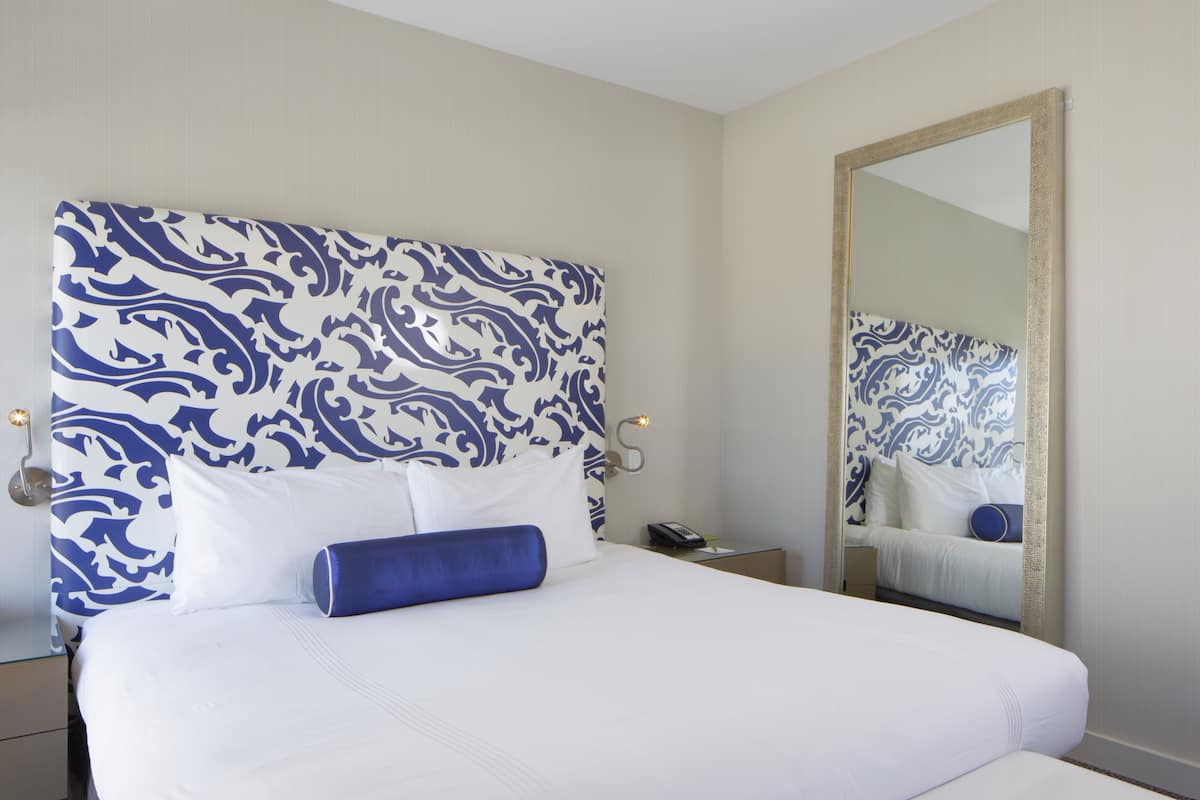 A hotel room with a large bed, a headboard with a blue and white pattern, a bedside table with a phone, and a mirror on the wall.